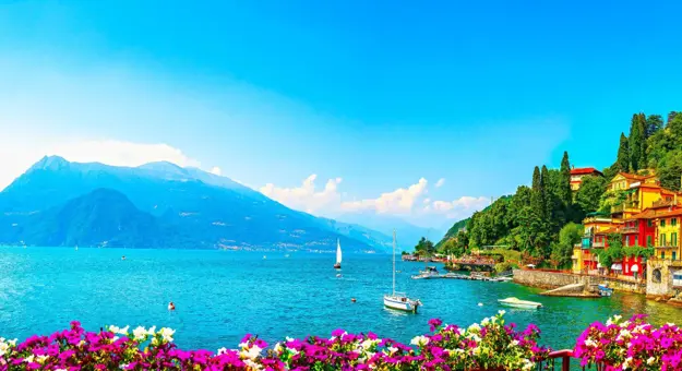 Colourful hillside houses in Lake Como overlooking the lake, where a couple of boats gently float, with mountains in the background