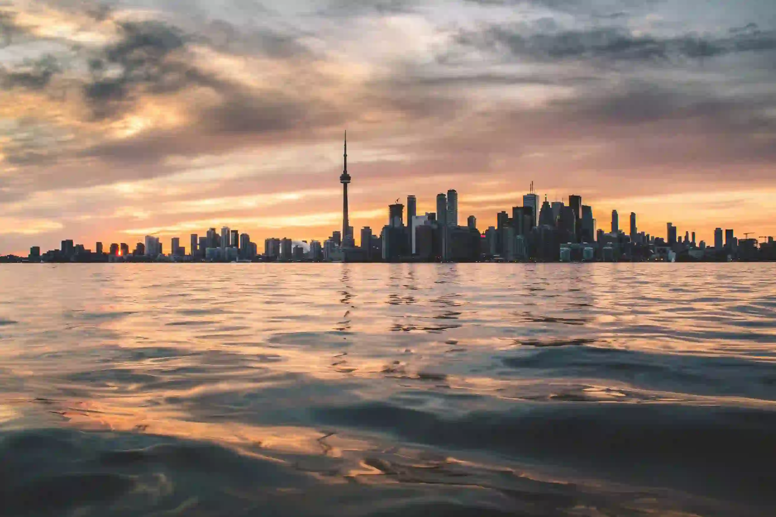 View over the water towards the Toronto skyline at sunset, with the CN Tower and surrounding buildings silhouetted against a pink and orange sky