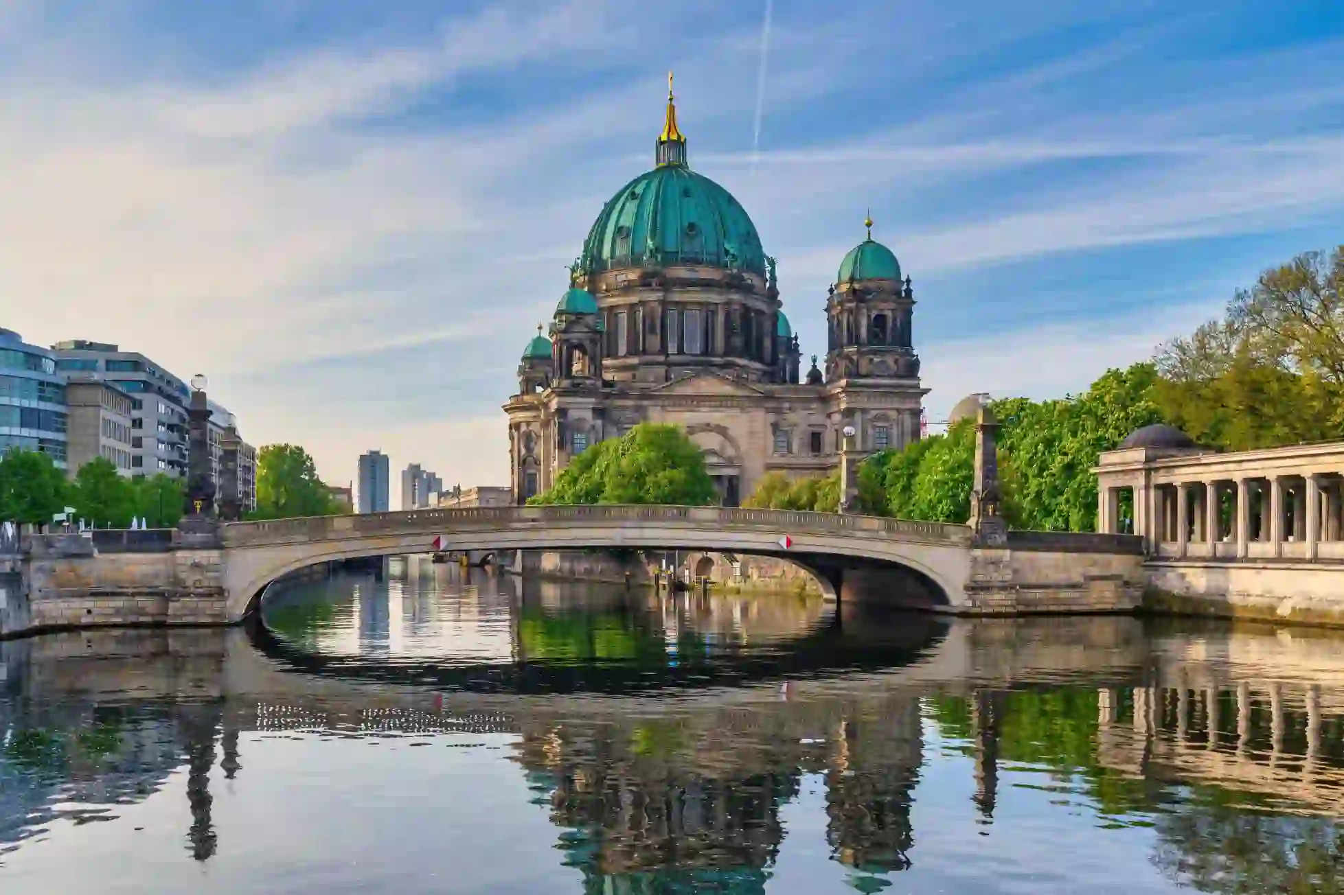 View of the Berlin Cathedral (Berliner Dom) with its green dome reflected in the River Spree, framed by a stone bridge, historic colonnades, and modern city buildings under a lightly clouded sky