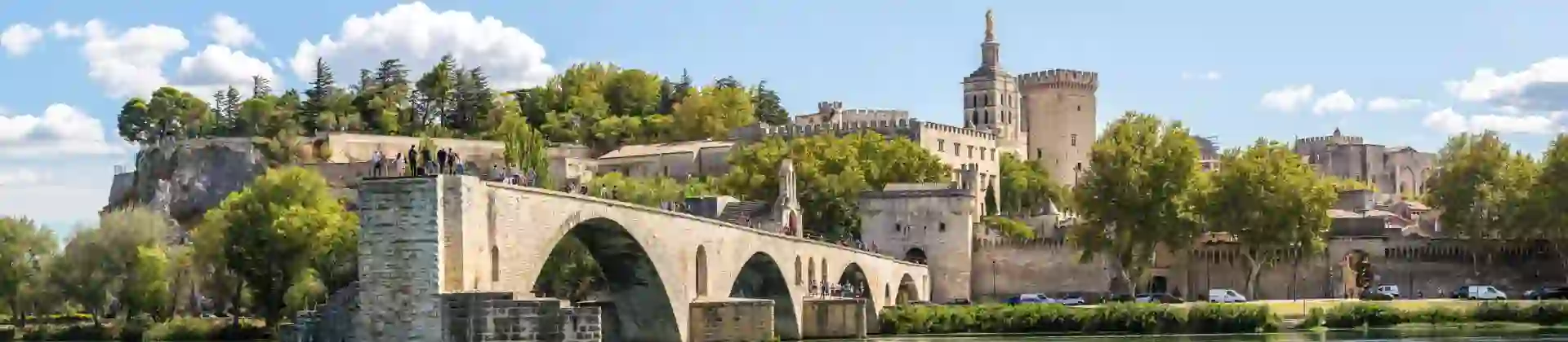Wide angle shot of the Pont d'Avignon bridge, with the palace on the other end. The river water in the forefront, and a blue sky with clouds above.