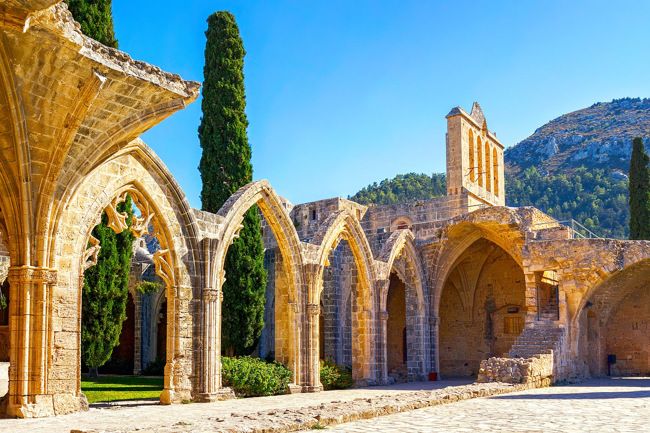 Low angle shot of the ruins of four brick archways connected to a wall with steps attached to it, and a thin, rectangular structure on top. Behind is the view of the mountain in front of a blue sky. To the left, behind the archway, two tall dark green trees.