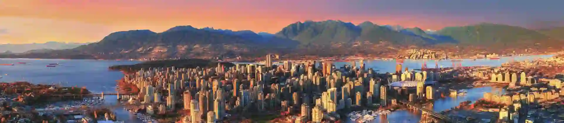 Aerial view of Vancouver, Canada at sunset, showing the city skyline, surrounding water, and distant mountains beneath a glowing, colourful sky