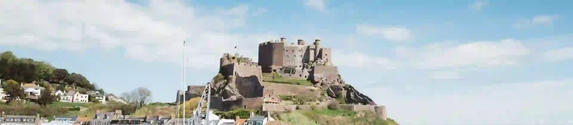 Long shot of Mont Orgueil Castle, showing part of the harbour below it and two people sat on the wall between the harbour and the sea, the buildings beside the harbour and the houses on a hill behind it, on a sunny day with a blue sky