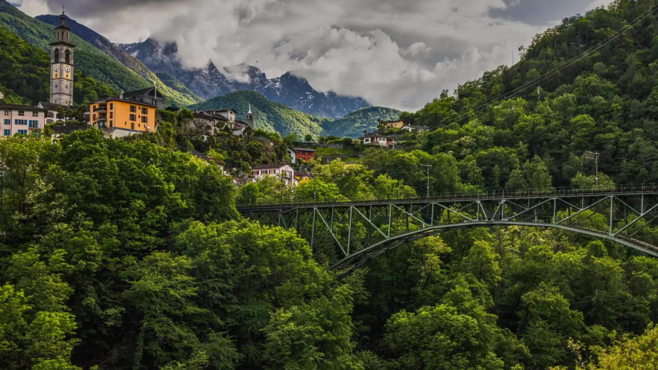 Landscape view of the Centovalli Railway bridge, surrounded by tall green trees, with mountains and a distant church tower in the background
