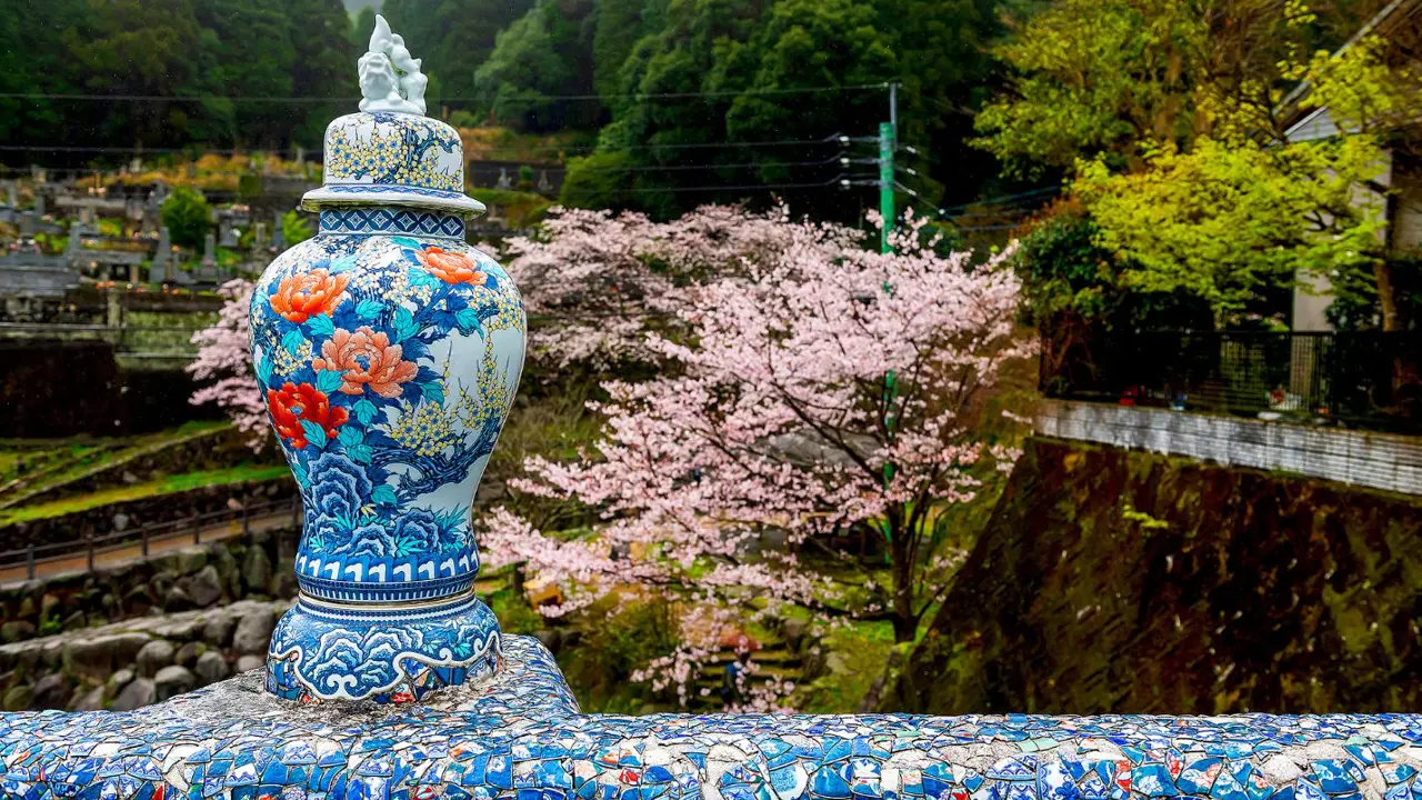 A decorative porcelain vase on a mosaic-tiled bridge in Okawachiyama village, with cherry blossoms and forested hills in the background