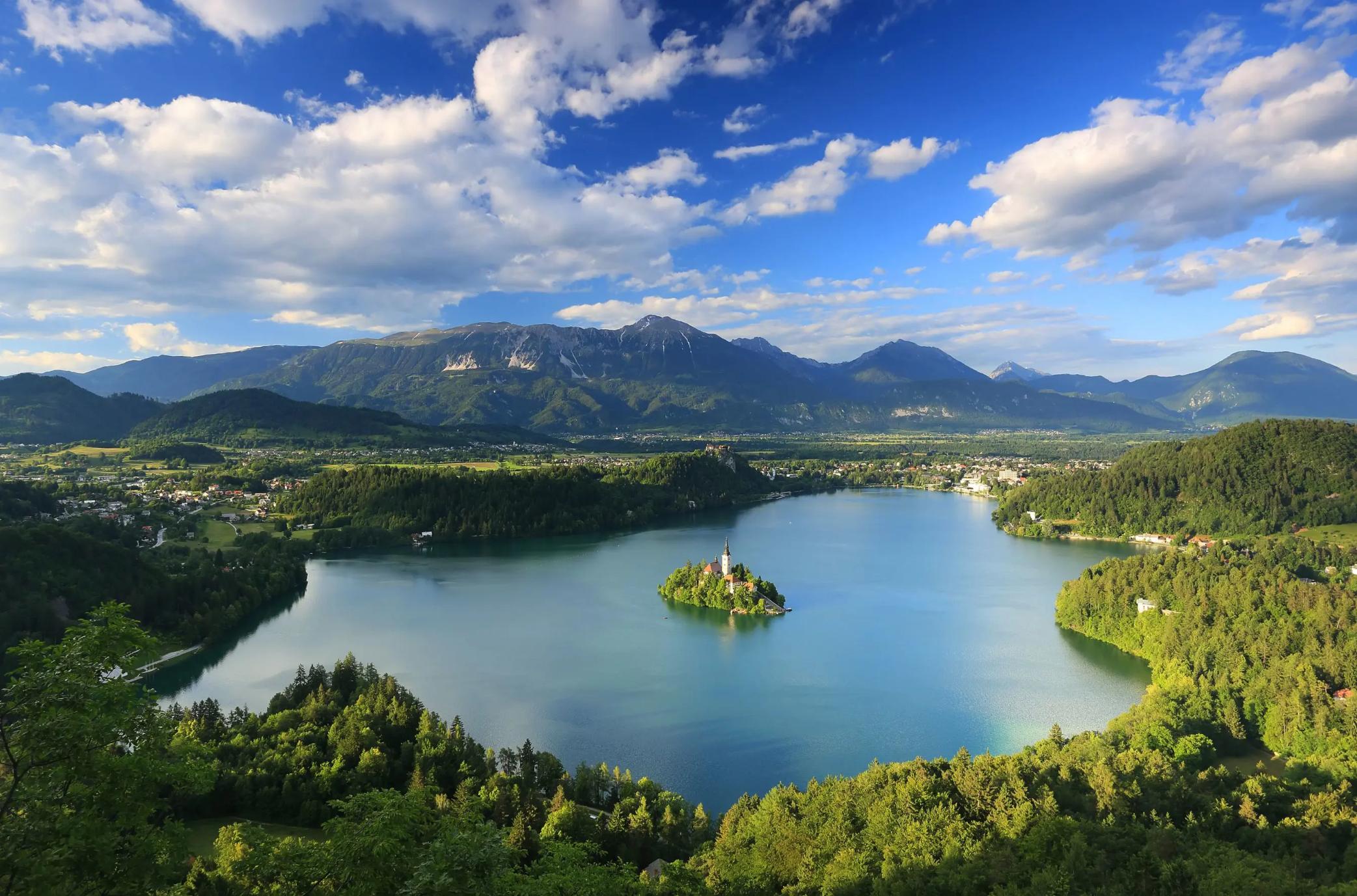 Wide angle shot of Lake Bled, showing its forested surrounding areas, and mountains in the distance. In the middle of the lake, there is a small island with trees and a towered building