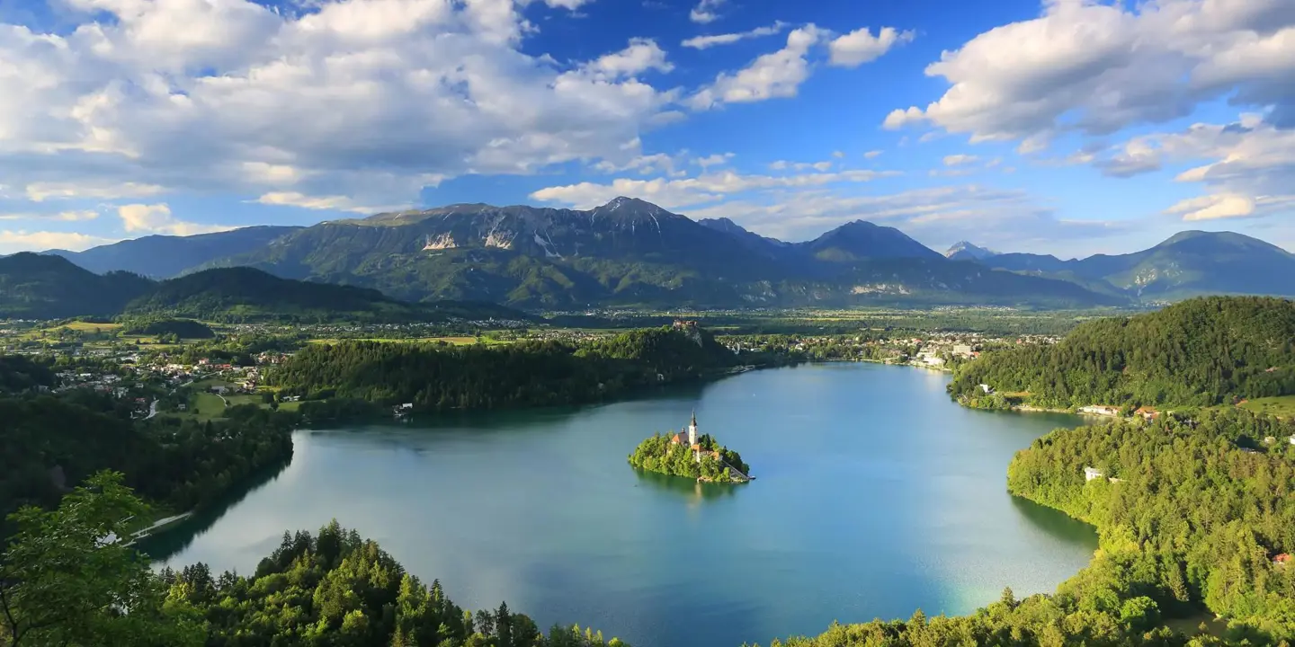 Wide angle shot of Lake Bled, showing its forested surrounding areas, and mountains in the distance. In the middle of the lake, there is a small island with trees and a towered building