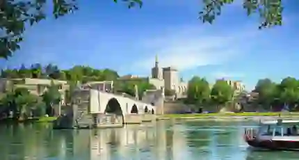View of the Pont D’Avignon bridge, with the river water in the forefront and a boat coming from the right. In the distance, on the other side of the bridge is a grey palace. The sky is blue.