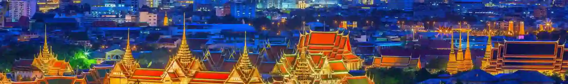 Distant view of the Grand Palace in Bangkok lit up at night, with its golden spires and ornate rooftops surrounded by softly illuminated temples and skyscrapers