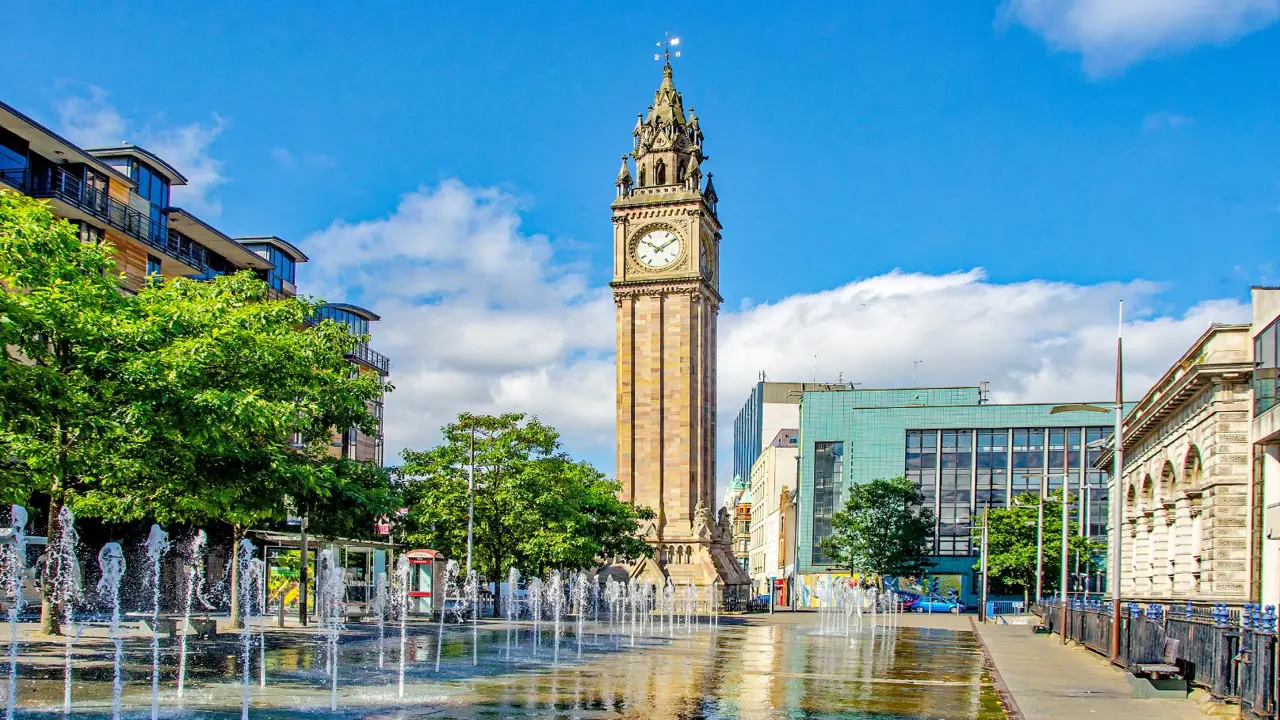 The Albert Memorial Clock tower rises above a plaza with fountains, surrounded by trees and modern city buildings under a blue sky