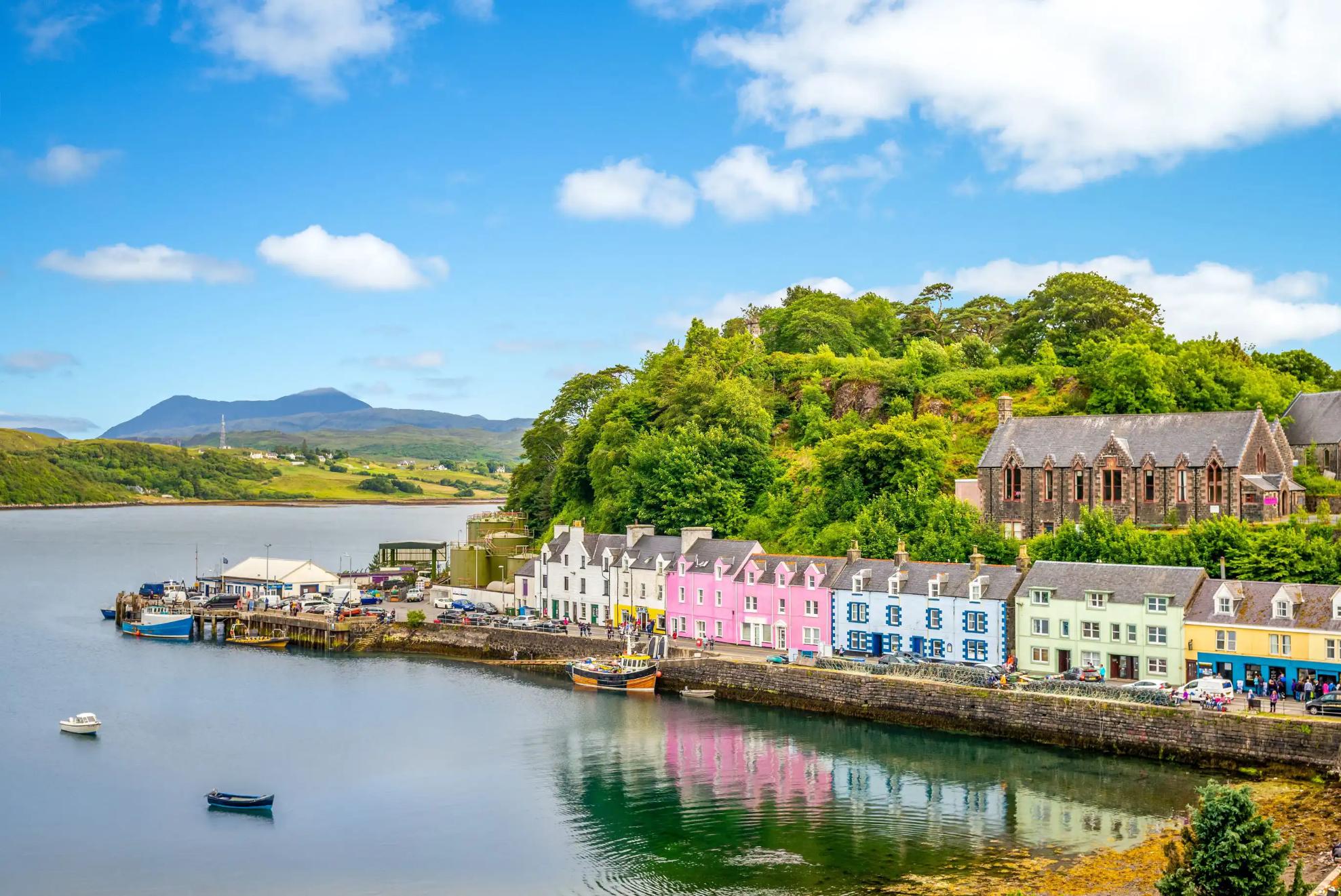 The colourful harbourfront of Portree on the Isle of Skye in Scotland, with pastel-painted houses reflected in the calm water, nestled beneath a lush green hill on a sunny day