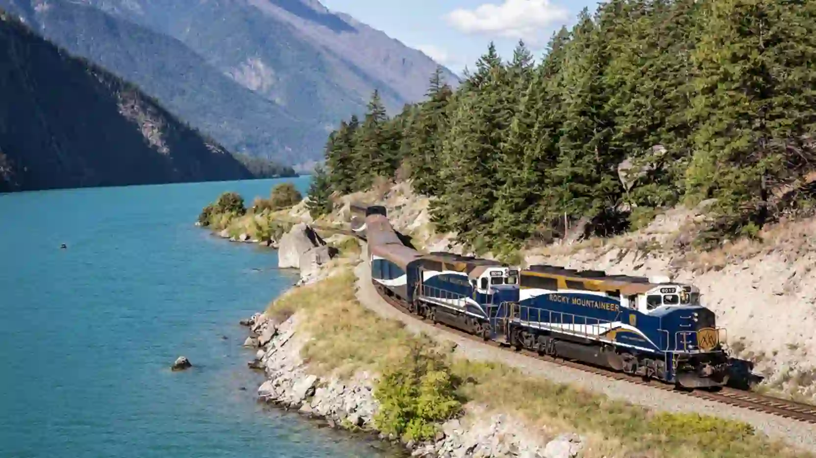 The Rocky Mountaineer train travelling through Canada beside a tranquil lake, with tall trees and scenic mountain views in the background