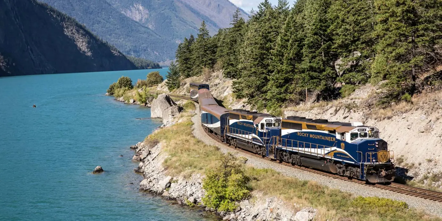 The Rocky Mountaineer train travelling through Canada beside a tranquil lake, with tall trees and scenic mountain views in the background
