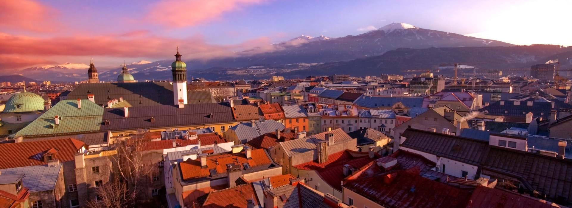 High angle shot of the city of Innsbruck, showing many roofs of houses and other buildings, with a few towers poking out, and one with a dome-shaped roof. Large mountains in the distance, dark pink clouds to the left and a blue sky. 