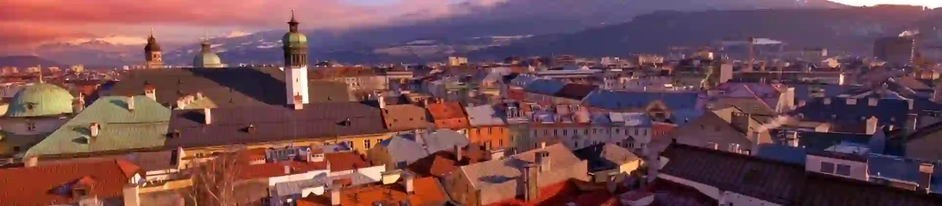 High angle shot of the city of Innsbruck, showing many roofs of houses and other buildings, with a few towers poking out, and one with a dome-shaped roof. Large mountains in the distance, dark pink clouds to the left and a blue sky.