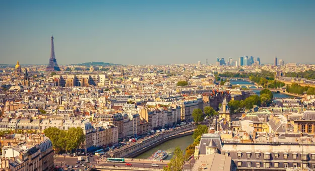 High angle view of the city of Paris, with a bridge and a main road with cars in the forefront. View of all the buildings of the city, with the Eiffel tower poking out in the distance to the left. To the right, behind the river, the faint view of skyscrapers in the distance.