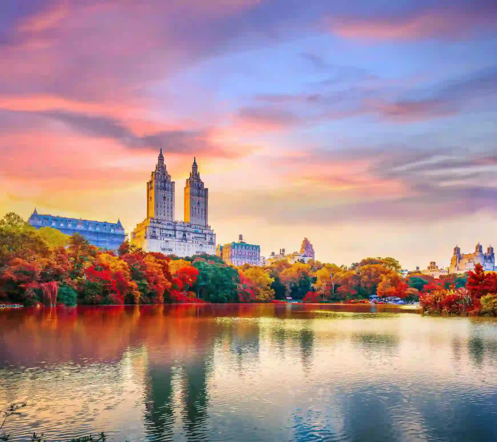 Central Park in autumn with The Lake in the centre, surrounded by colourful trees in shades of red, orange, green and yellow. The iconic twin towers of The San Remo rise in the background