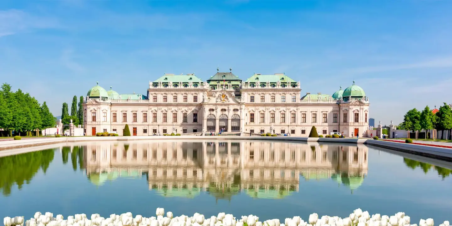 The Belvedere Palace in Vienna, Austria, reflected in a large pond with white tulips and other flowers in the foreground under a clear blue sky