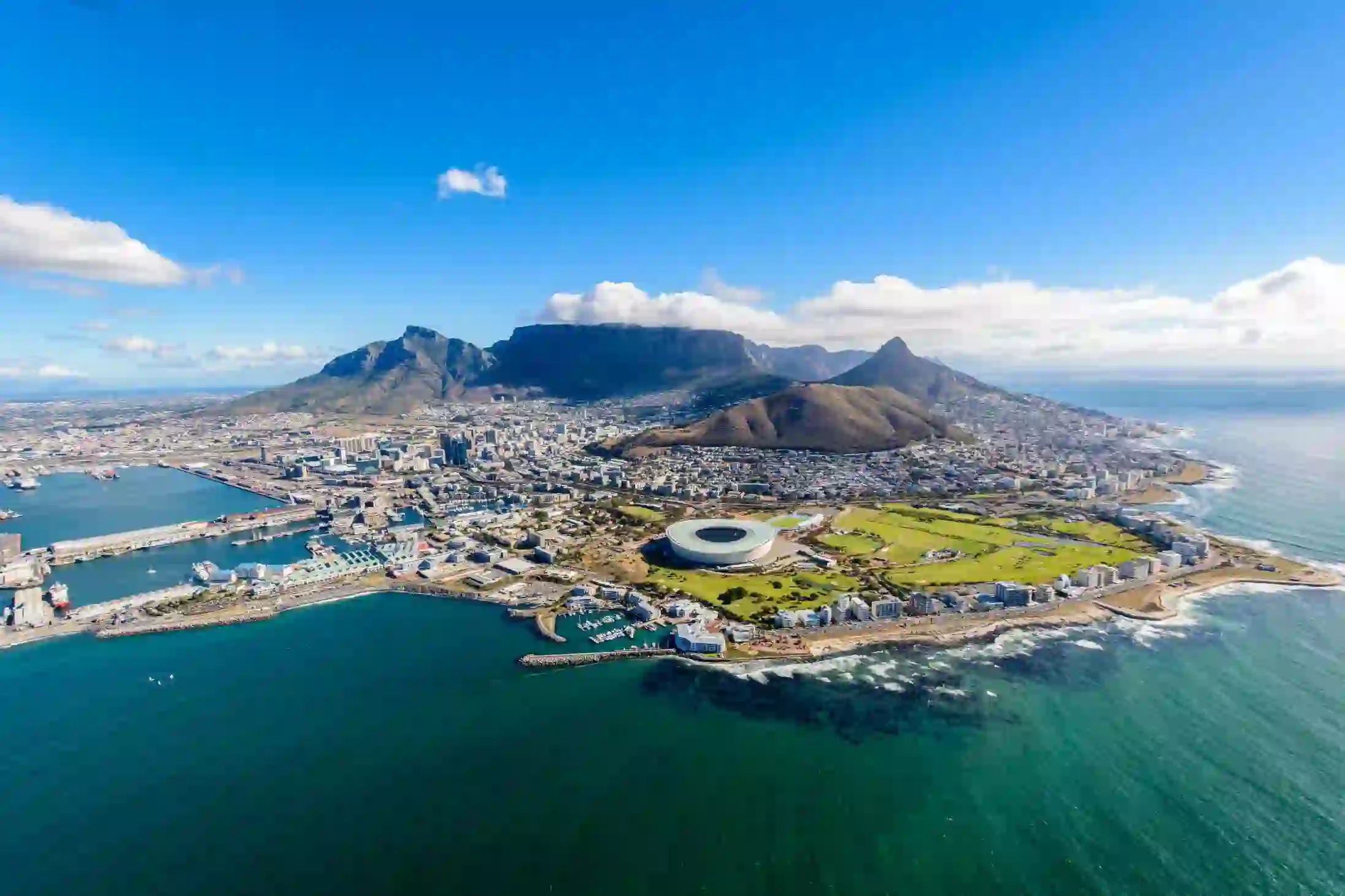 Aerial view of Cape Town, South Africa, showing the city nestled between Table Mountain and the Atlantic Ocean, with the coastline, harbour, and urban landscape clearly visible