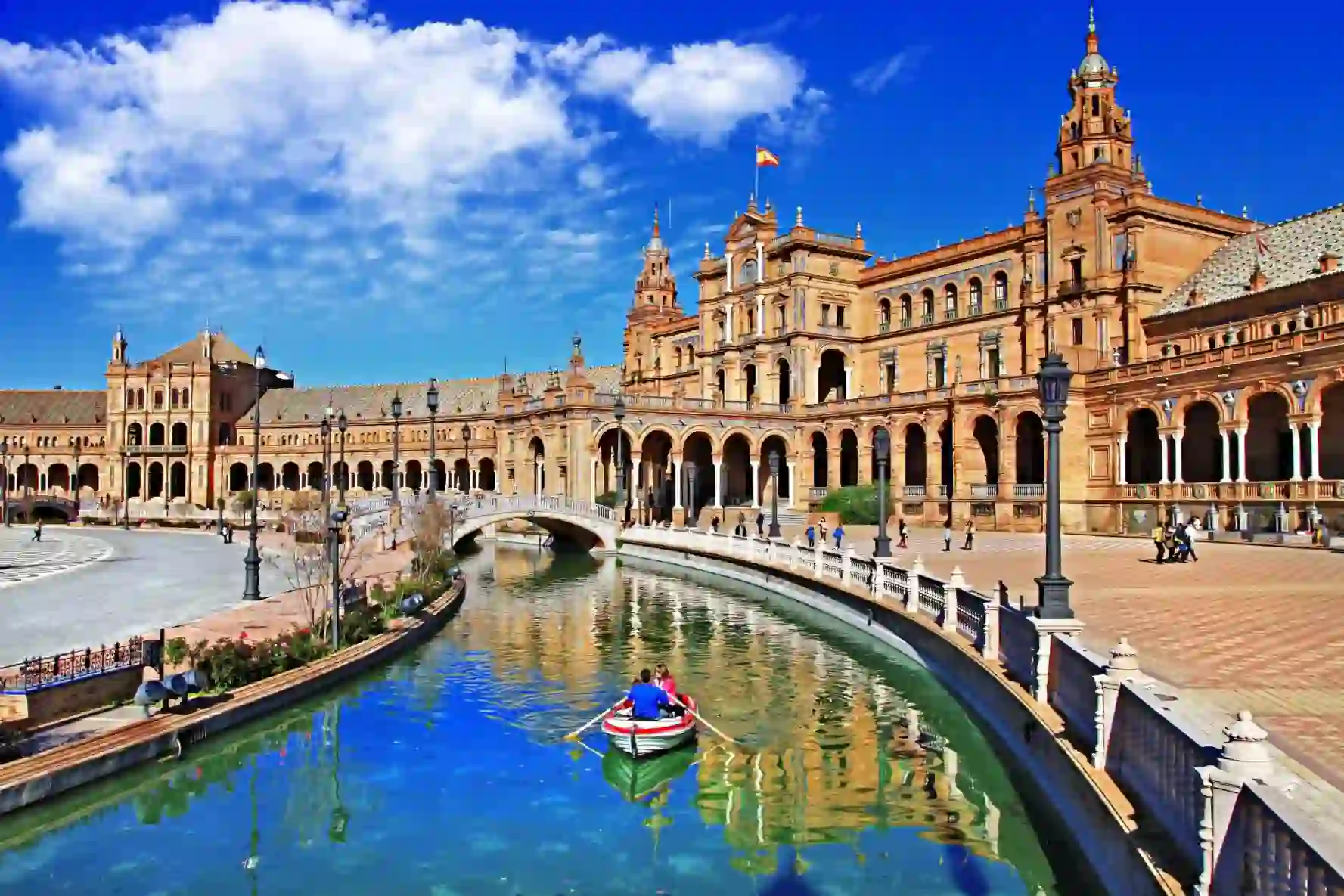 A couple rows a boat along the canal at Plaza de España in Seville, Spain, with the historic buildings and a bright blue sky reflected in the water