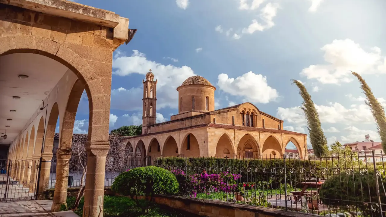 A stone Greek Orthodox monastery with arched walkways, a domed roof, and a bell tower. A well-kept garden with flowers and shrubs lines the foreground under a sunny sky