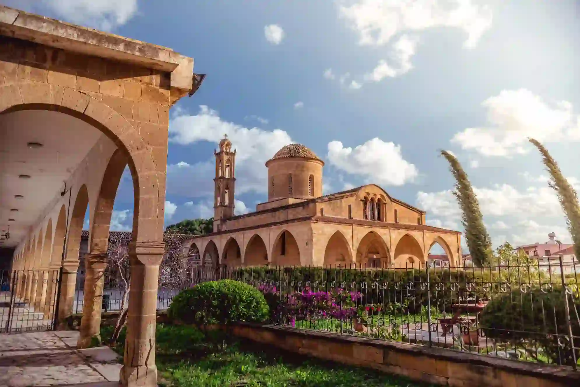 A stone Greek Orthodox monastery with arched walkways, a domed roof, and a bell tower. A well-kept garden with flowers and shrubs lines the foreground under a sunny sky