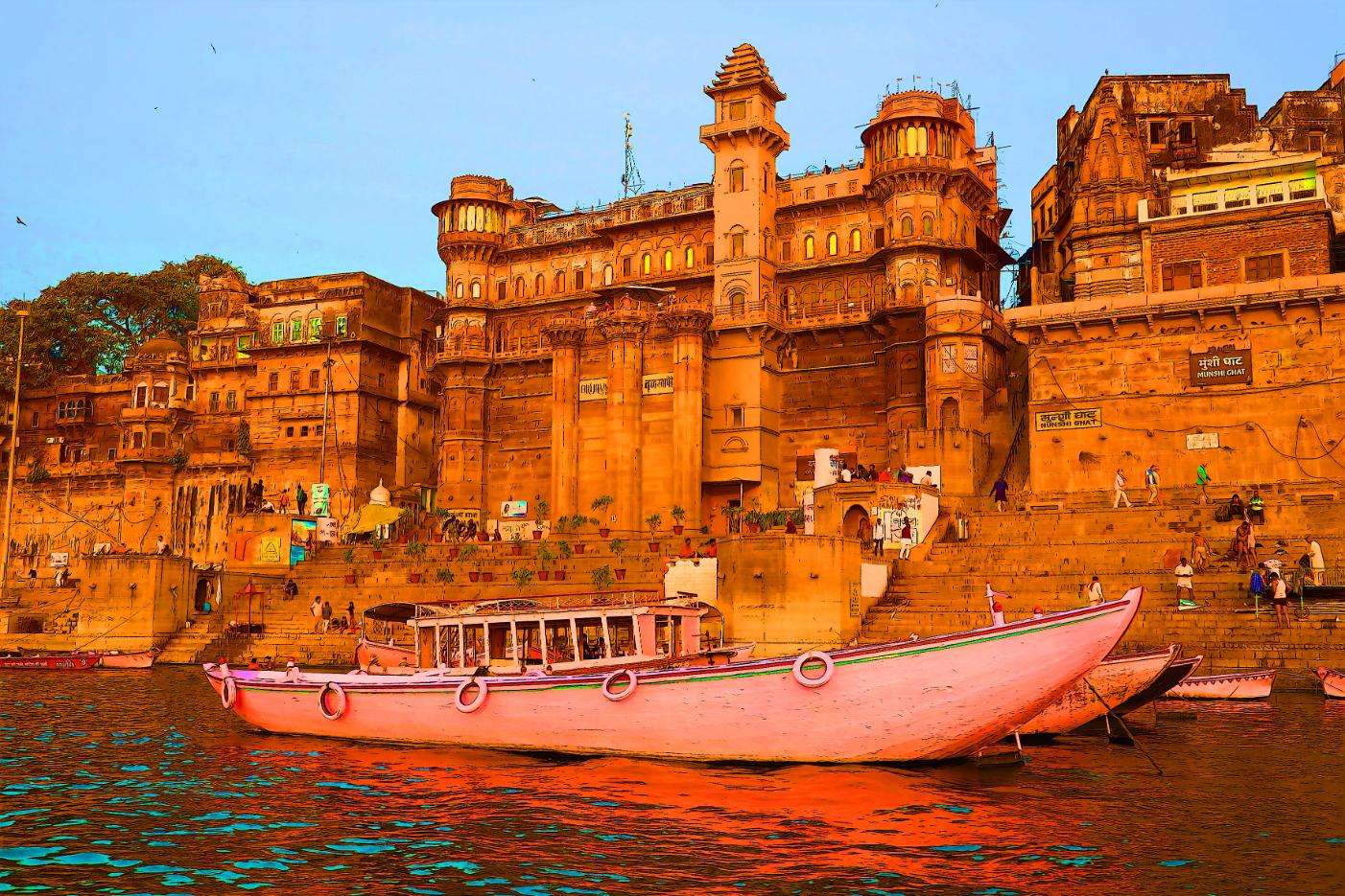 The ghats of Varanasi seen from the River Ganges, with historic sandstone palaces and colourful boats reflecting in the calm evening water
