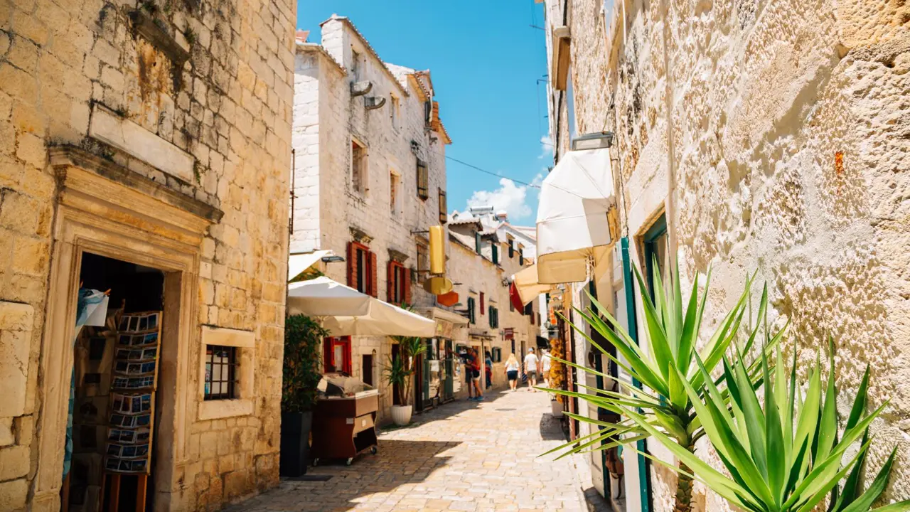 A charming narrow alleyway in Split’s Old Town lined with old stone buildings, red-shuttered windows, hanging signs, and small shops, with potted plants and tourists strolling beneath a clear blue sky