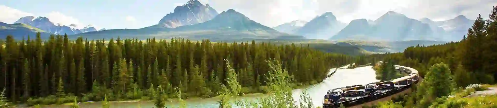 The Rocky Mountaineer train travelling through the Canadian Rockies, with rugged mountains in the background and dense evergreen forests lining the tracks