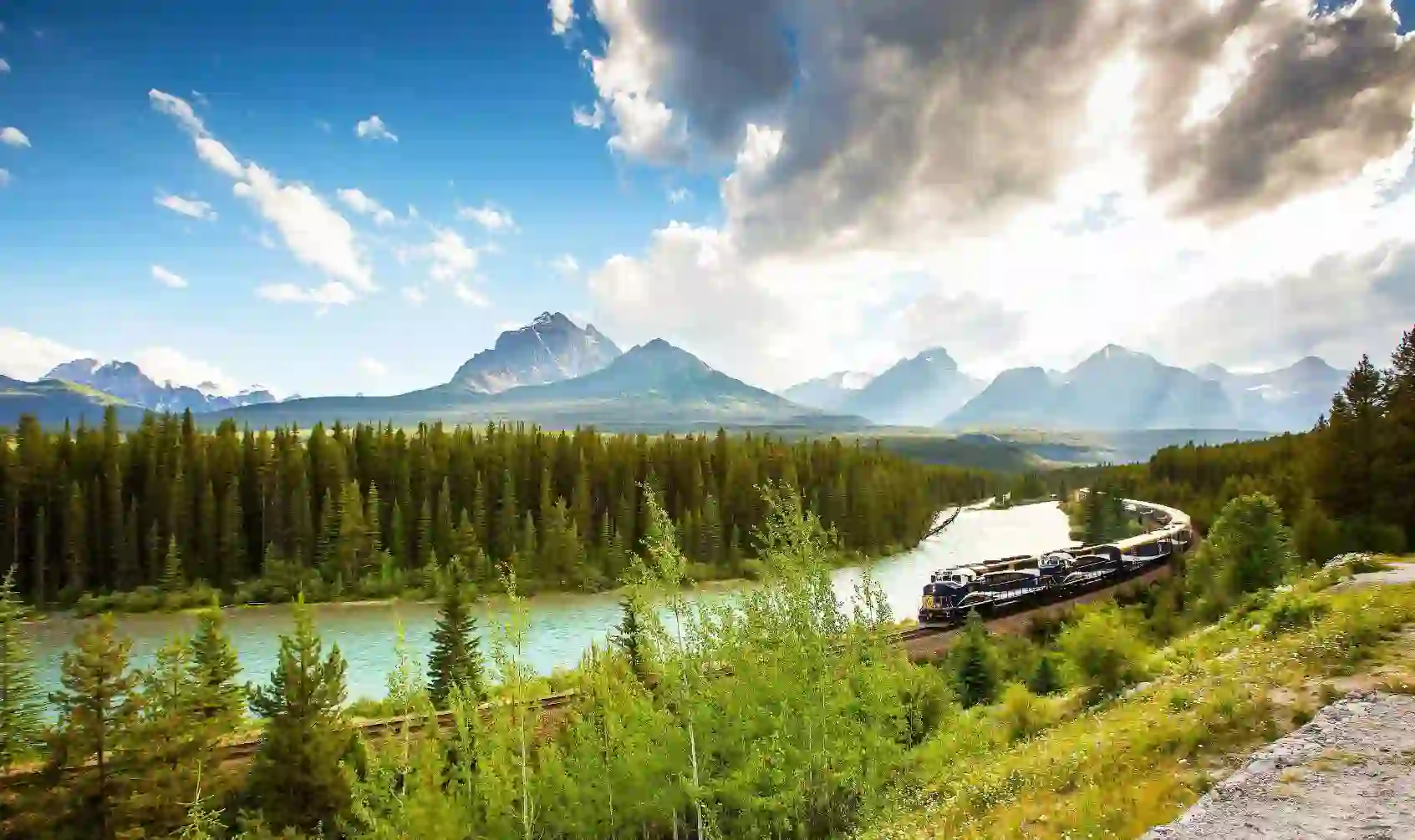 The Rocky Mountaineer train travelling through the Canadian Rockies, with rugged mountains in the background and dense evergreen forests lining the tracks