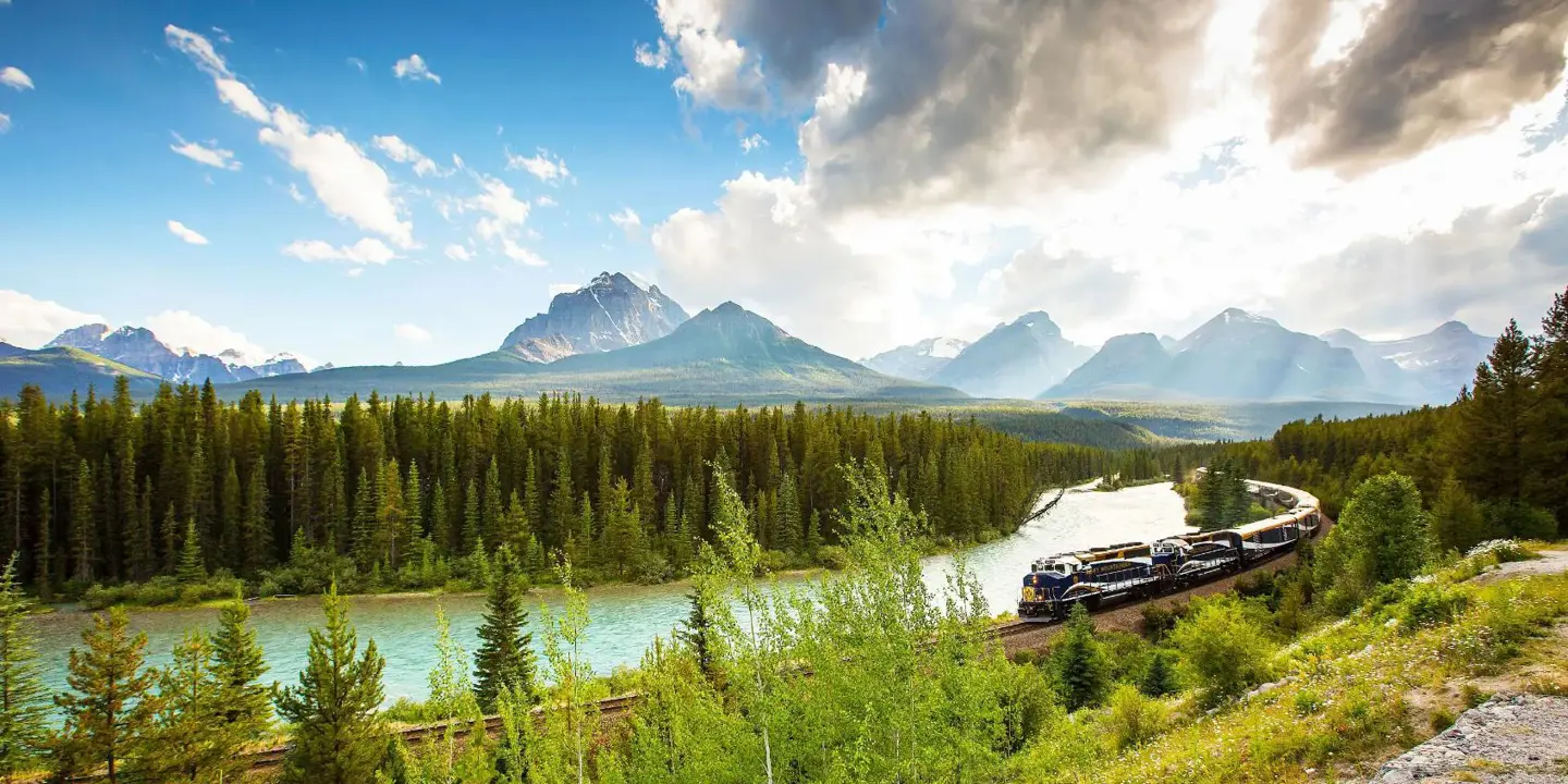 The Rocky Mountaineer train travelling through the Canadian Rockies, with rugged mountains in the background and dense evergreen forests lining the tracks